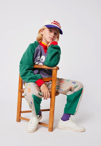 Outfit colorato per bambini che include un felpa verde e grigia, pantaloni beige a fantasia e scarpe bianche, seduto su una sedia di legno.