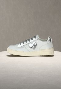 Λευκό αθλητικό παπούτσι χαμηλής κοπής με ασημένια κορδόνια και τακούνι, διακοσμημένο με ασημένια καρδιά και το κείμενο "LOVE MOSCHINO" στο πλάι.