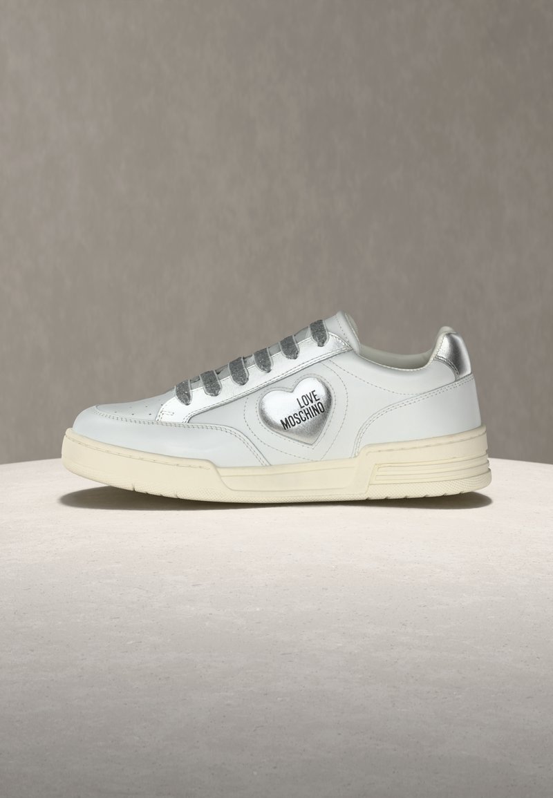 Λευκό αθλητικό παπούτσι χαμηλής κοπής με ασημένια κορδόνια και τακούνι, διακοσμημένο με ασημένια καρδιά και το κείμενο "LOVE MOSCHINO" στο πλάι.