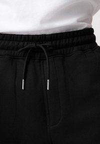 Gros plan sur la taille d'un pantalon de jogging noir avec élastique et cordon noué, porté avec une chemise blanche rentrée.