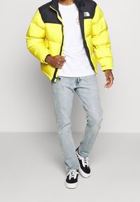 Żółta kurtka puchowa z czarnymi akcentami, z zamkiem błyskawicznym i logo na lewej piersi, noszona na białym t-shircie i jasnoniebieskich dżinsach.