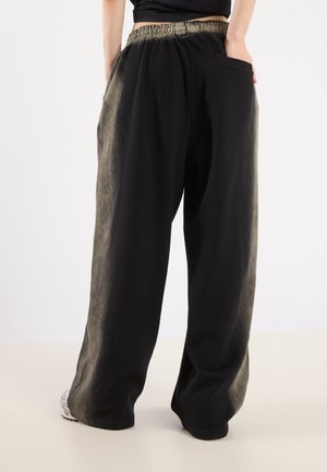 Person wearing loose black pants with elastic waistband and side pockets, standing with one hand in pocket, white sneakers visible.