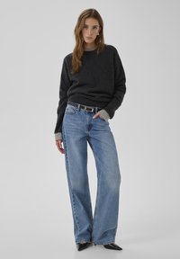 Dunkelgrauer Strickpullover mit überschnittenen Schultern, kombiniert mit hellblauen, weit geschnittenen Jeans. Schwarze spitze Schuhe und ein schwarzer Gürtel.