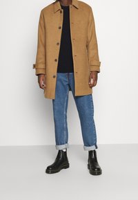 Tan ullrock med krage, knappstängning och uppvikta ärmar. Bärs över en marinblå tröja och blå jeans. Svarta ankelstövlar fulländar outfiten.