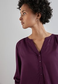 Blusa viola con scollatura a V, dettaglio di bottoni e maniche lunghe. Il tessuto appare liscio con una morbida texture e una vestibilità rilassata.