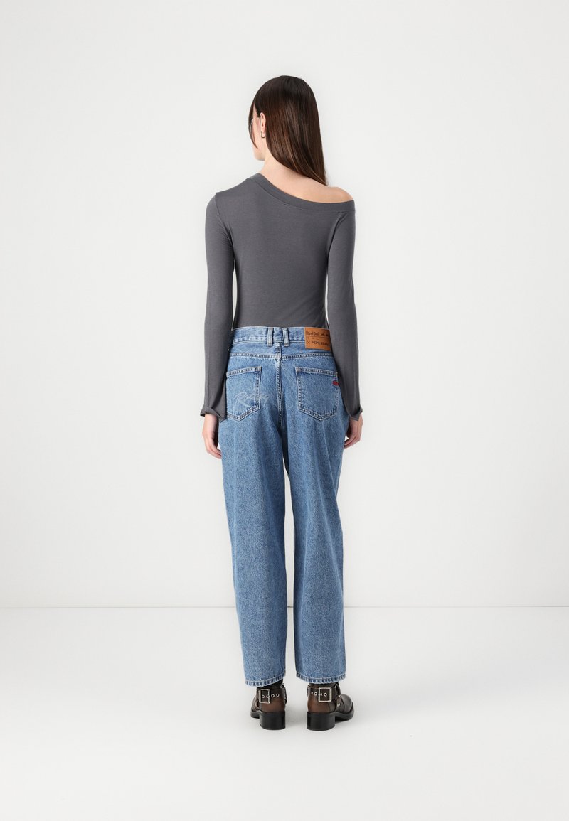 Graues Langarmoberteil mit asymmetrischem Ausschnitt, kombiniert mit lockeren blauen Jeans. Auffällige Stickereien an den Hintertaschen und ein braunes Lederetikett.