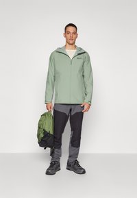 Chaqueta impermeable verde claro con capucha, combinada con pantalones grises y negros. Se sostiene una mochila verde con detalles negros en una mano.