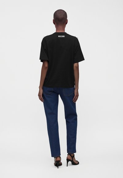 Personne portant un t-shirt noir Moschino, un jean bleu et des sandales noires à talons hauts, debout dos à la caméra.