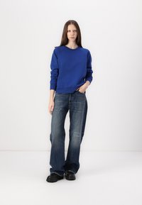 Felpa blu con testo in rilievo, abbinata a jeans denim scuri a vestibilità comoda. Scarpe nere chunky. Sfondo bianco semplice.