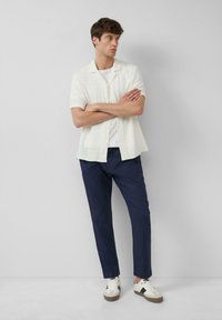 Korte mouwen witte linnen shirt, marineblauwe broek, witte sneakers met bruine accenten. Model staat met de armen gekruist tegen een eenvoudige achtergrond.