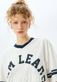 Hvid oversized T-shirt med en marineblå V-hals, prydet med store sorte bokstaver, der siger "CALIFORNIA." Blødt bomuldsstof med korte ærmer.