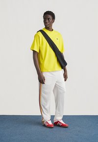 Άνδρας με κίτρινο πουκάμισο Adidas, λευκό παντελόνι με πολύχρωμες ρίγες, κόκκινα αθλητικά παπούτσια και μαύρη τσάντα χιαστί, σε μπλε πάτωμα μπροστά σε λευκό τοίχο.