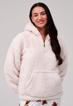 Jeune femme aux longs cheveux foncés portant un sweat à capuche duveteux rose clair zippé et un pantalon blanc avec des motifs d'oursons, souriant doucement.