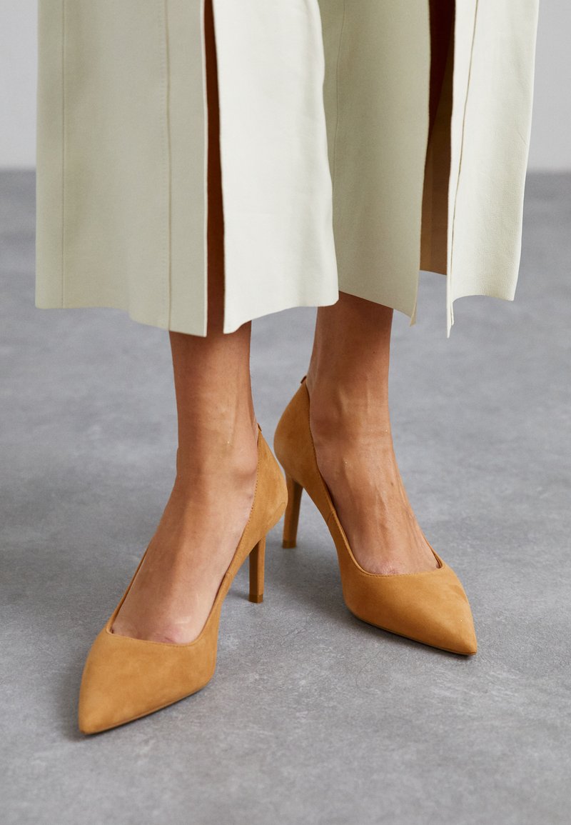 Escarpins en daim beige à talons hauts avec un bout pointu et des côtés découpés, associés à un pantalon clair ajusté avec des fentes sur les côtés.