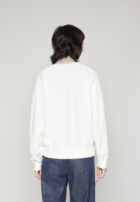 Witte sweatshirt met een relaxte pasvorm, geribde manchetten en zoom, en een zachte textuur. Draagt met blauwe spijkerbroek, gezien vanachter.