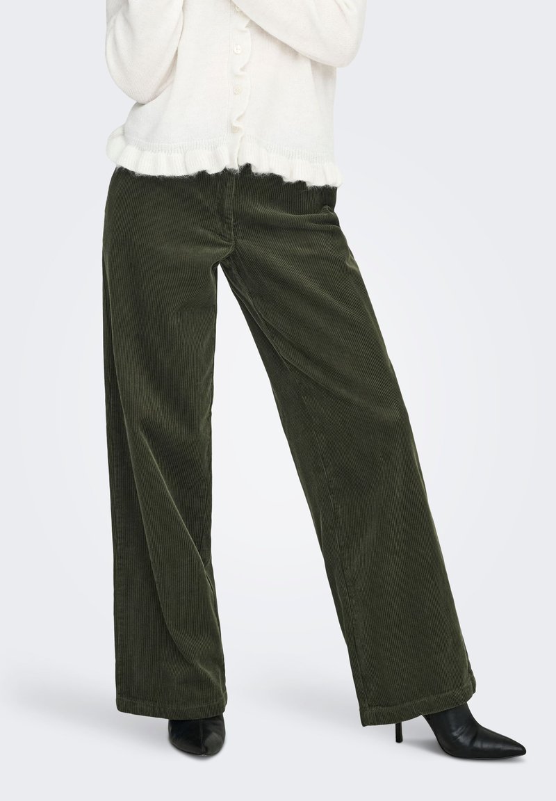 Femme portant un pantalon en velours côtelé vert foncé à jambes larges et des bottines noires à bout pointu, avec un pull blanc texturé boutonné.