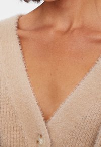 Béžový, strukturovaný kardigan s designem do V a měkkou, chlupatou látkou. Obsahuje zapínání na knoflík u krku. Jemně žebrovaný vzor.