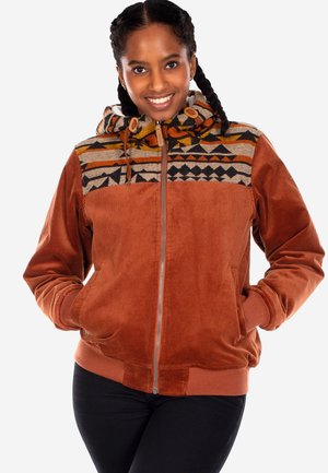 Young woman with braided hair smiles wearing a rust-colored zip-up jacket with a patterned hood and black pants.