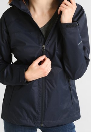 Persoon draagt een marineblauwe jas met een gedeeltelijk dichtgeritste voorkant, die met de rechterhand de kraag aanpast.