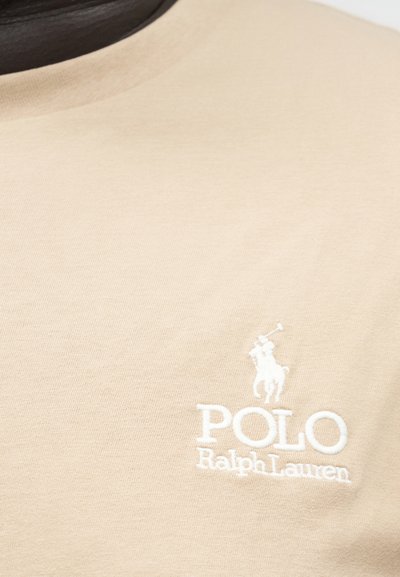 Beige stoff med hvit brodert Polo Ralph Lauren-logo som viser en polo-spiller til hest med en kølle.