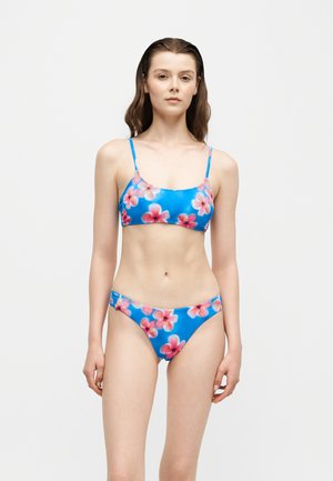 Młoda kobieta w niebieskim bikini z różowym kwiatowym wzorem stoi na jednolitym białym tle.