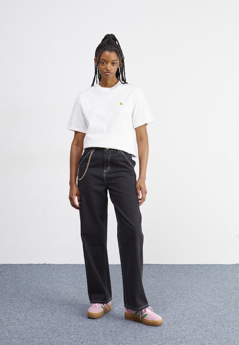 Hvid bomuldst-shirt med et lille gult logo, kombineret med mørke denimbukser med en kædedetalje. Lyserøde platform-sneakers med grønne accenter.