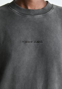 Felpa grigia realizzata in un morbido tessuto testurizzato, con un colletto rotondo a coste e logo "TOMMY JEANS" in rilievo sulla parte anteriore.