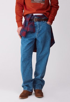 Mężczyzna w luźnych niebieskich dżinsach, brązowym pasku, czerwonej kraciastej koszuli zawiązanej w talii, bluzie w kolorze spalonym pomarańczowym oraz brązowych zamszowych butach.