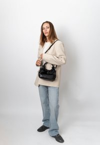 Femme portant un blazer beige, un jean bleu et des chaussures noires, tenant un sac à main noir avec des détails froncés sur fond blanc.