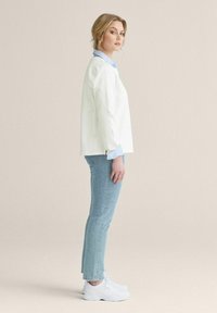 Vit långärmad topp över en ljusblå skjorta med krage, tillsammans med ljusblå jeans och vita sneakers. Sidovy från en neutral bakgrund.