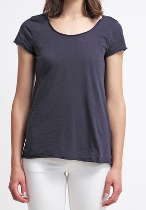 Kobieta ubrana w ciemnoniebieski t-shirt z krótkim rękawem i dekoltem w serek oraz białe spodnie, stojąca ze swobodnie opuszczonymi ramionami na tle jednolitego tła.