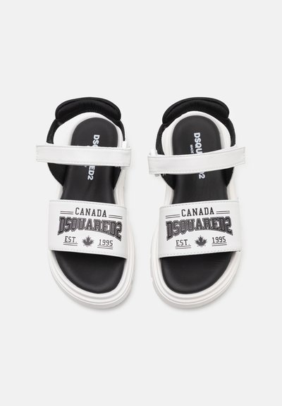 Bela sandala z črnimi poudarki, z nastavljivimi trakovi, oblazinjenim vložkom in boldnim napisom "Dsquared2 Canada" ter logotipom.