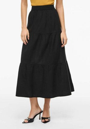 Kvinne iført et svart lagdelt midiskjørt med elastisk midjebånd og svarte åpne hælsandaler.