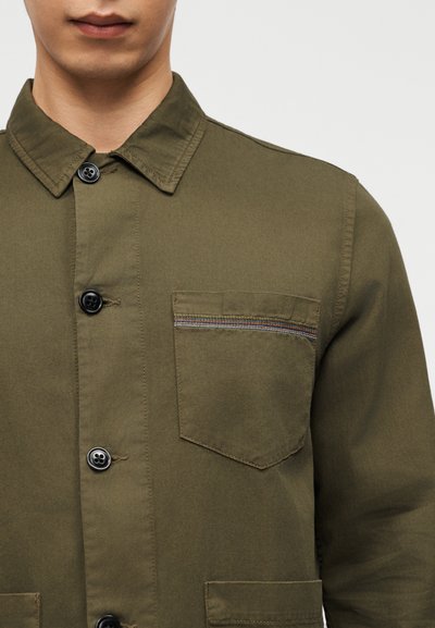 Άνδρας που φοράει πράσινο-ελαιολάδι σακάκι με κουμπιά και γιακά, με μια τσέπη στο στήθος που έχει λεπτές πολύχρωμες ρίγες κοντά στην πάνω άκρη.