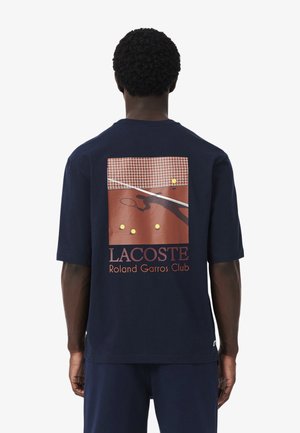 Uomo che indossa una maglietta Lacoste blu navy con stampa di campo da tennis, rete, palline e ombra, con testo "Roland Garros Club" sul retro.
