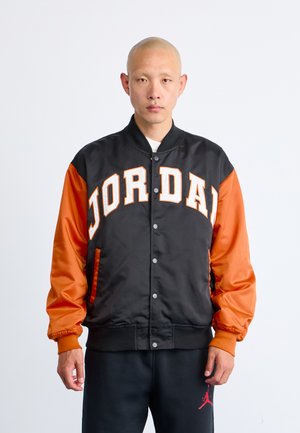Person wearing a black and orange varsity jacket with "JORDAN" in large letters across the chest and black pants with a red logo.