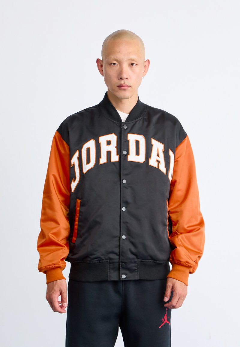 Person wearing a black and orange varsity jacket with "JORDAN" in large letters across the chest and black pants with a red logo.