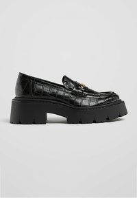 Mocassins noirs à motif crocodile avec une semelle épaisse noire et des accessoires en métal doré sur le dessus, présentant un design élégant et une finition brillante.