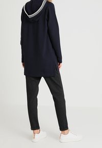 Sudadera azul marino con capucha a rayas, parte trasera larga, aberturas laterales, combinada con joggers oscuros de textura y zapatillas blancas.