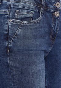 Close-up of dark blue denim jeans showing front pocket, coin pocket, and metal button details with contrast stitching.