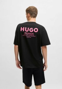 Henkilö, joka on pukeutunut mustiin shortseihin ja mustaan T-paitaan, jossa on vaaleanpunainen "HUGO" etupuolella ja teksti selässä, seisoo yksinkertaista vaaleaa taustaa vasten.