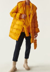 Abrigo acolchado amarillo con cremallera frontal y capucha, combinado con un suéter naranja, pantalones navy y botas de tobillo beige. Presenta tejido acolchado.