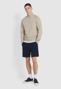 Jongeman staat met zijn handen in zijn zakken, gekleed in een beige sweatshirt met kwartrits, marineblauwe textuur shorts, witte sokken en marineblauwe sneakers op een witte achtergrond.