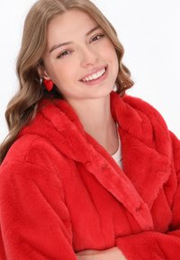 Giovane donna sorridente, che indossa un vivace cappotto di pelliccia sintetica rossa e orecchini a forma di cuore rossi, con capelli castano chiaro ondulati.