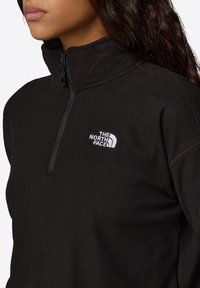 Czarny polarowy rozpinany sweter z wysokim kołnierzem, zapinany na zamek błyskawiczny do połowy oraz białym haftowanym logo po lewej stronie klatki piersiowej. Miękka tekstura i casualowy design.