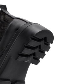 Zapatilla negra con parte superior de cuero texturizado y suela de goma resistente con un patrón en zigzag para mejorar la tracción. Primer plano del área del talón.