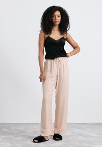 Vrouw met krullend haar die een zwarte kant-modieuze camisole, lichtroze wijde broeken en zwarte fluffy slippers draagt, staande met haar hand op de heup.