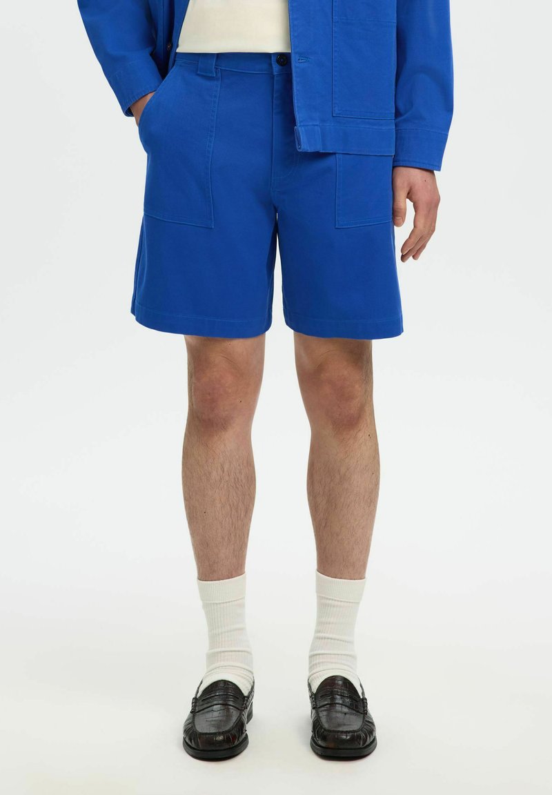 Personne portant un short bleu vif et une veste assortie, des chaussettes blanches, des mocassins noirs, debout devant un fond clair uni.