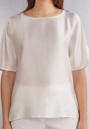 Donna che indossa una camicetta liscia color crema, manica corta, in raso, con scollatura rotonda e vestibilità comoda.