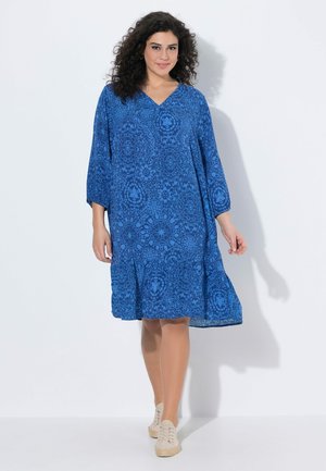 Frau mit lockigem Haar, die ein lockeres blau gemustertes knielanges Kleid und beige Sneakers trägt, steht vor weißem Hintergrund.